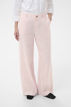 Femme portant un pantalon large en lin rose clair avec bouton sur le devant et poches, associé à une chemise blanche et des chaussures noires.