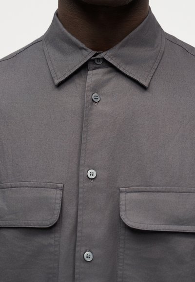Σκούρο γκρι πουκάμισο με κουμπιά και δύο τσέπες στο στήθος με καπάκι, φορεμένο από άτομο με σκουρόχρωμο δέρμα, εμφανιζόμενο από τον λαιμό έως το μέσο του κορμού.
