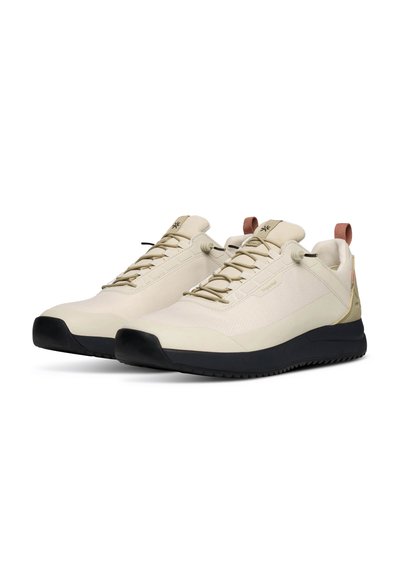 Paire de baskets basses beige avec semelles noires, lacets élastiques et une languette de tirage beige sur le talon, présentées légèrement en angle sur fond blanc.