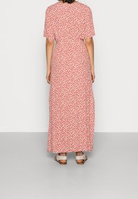 Maxi sukienka w kolorze koralowym z białym wzorem liści, krótkie rękawy i dopasowana talia. Wykonana z lekkiego materiału. Sandały widoczne na stopach.