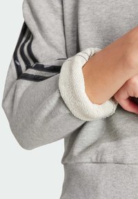 Sudadera gris con puños acanalados en una textura de tejido contrastante. Incluye rayas negras en la manga para un detalle y diseño adicionales.