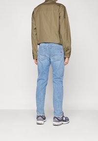 Camisa verde oliva de manga larga, jeans de mezclilla azul claros y zapatillas deportivas grises. Cuenta con bolsillos estándar y un corte holgado.