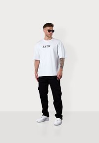 Weißes Oversized-T-Shirt mit schwarzem "ESTM"-Druck, kombiniert mit schwarzen Cargo-Hosen und weißen Sneakers. Das Model trägt eine dunkle Sonnenbrille.