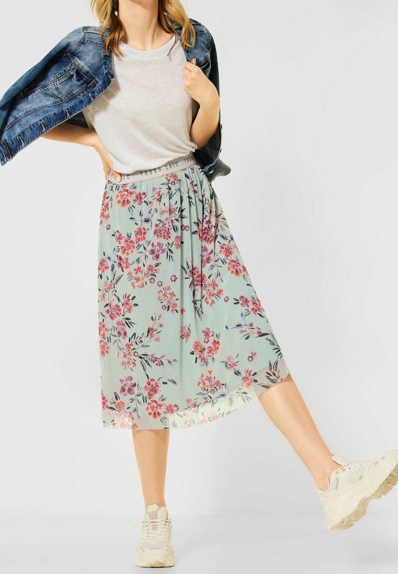 Falda midi floral en verde claro, con flores rosas y moradas, cintura elástica y caída suave. Combinada con una chaqueta vaquera y zapatillas.