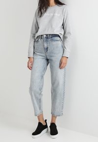 Felpa grigia con la scritta "CALVIN KLEIN JEANS", jeans a vita alta blu chiaro e scarpe slip-on nere con suole bianche.