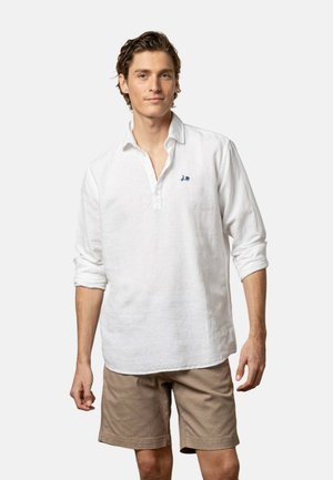 Joven con una camiseta polo blanca de manga larga con un pequeño logo azul y pantalones cortos beige hasta la rodilla, de pie frente a un fondo liso.