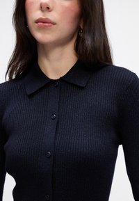 Námornícky žebrovaný cardigan s golierom, na gombíky a s jemným leskom. Tkanina je mäkká a vyrobená z ľahkého, elastického materiálu.