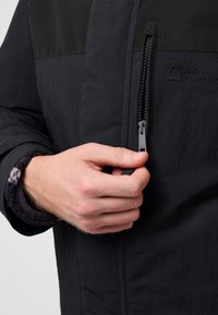 Schwarze Fleece-Jacke mit grauen Akzenten, ausgestattet mit einem Reißverschluss, strukturiertem Oberteil und einem Logo im Brustbereich.