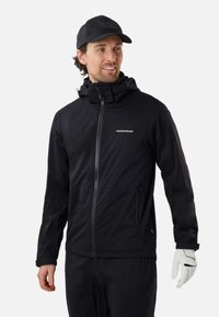 Homme portant une veste noire zippée avec le logo "Swedemount", casquette noire et gant de golf blanc à la main droite, regardant légèrement vers la droite.