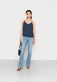 Top sem mangas azul-marinho com padrão floral branco, design drapeado, combinado com jeans claros de boca de sino e uma bolsa preta. Sandálias rasas usadas.
