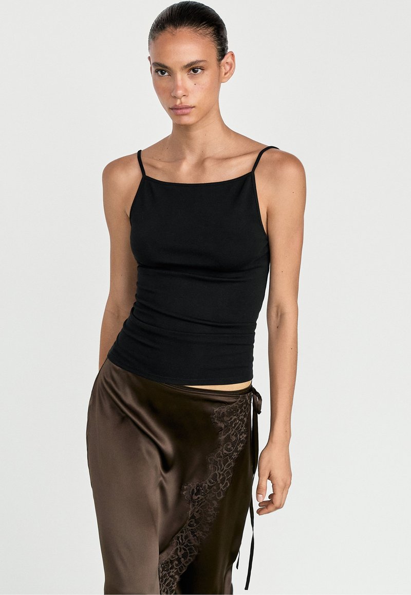 Woman wearing a black spaghetti strap top and a brown silk skirt with lace detail standing against a plain background.