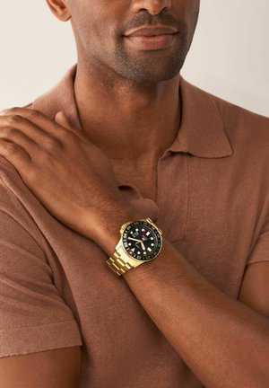 Homme portant une chemise marron à manches courtes avec une montre-bracelet dorée avec un cadran noir et un bracelet en métal, bras croisés sur la poitrine.
