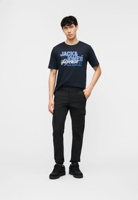 Uomo in piedi che indossa pantaloni cargo neri, scarpe da ginnastica nere e una maglietta blu navy con la scritta "Jack & Jones" in blu e bianco.