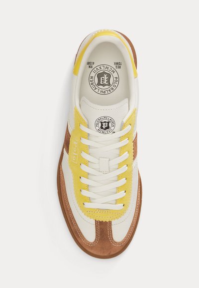 Άσπρο αθλητικό παπούτσι με κίτρινα και καφέ σουέντ στοιχεία, άσπρα κορδόνια και το λογότυπο Polo Ralph Lauren στη γλώσσα και την εσωτερική σόλα, ορατό από πάνω.