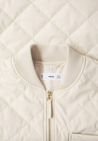 Veste matelassée crème avec col côtelé et fermeture éclair dorée, affichant l'étiquette de la marque "MNG" et les informations de taille à l'intérieur du col.