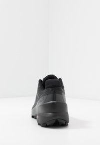 Chaussure de sport noire avec une tige en mesh texturé, un renfort au niveau du talon et une semelle en caoutchouc avec des pointes de traction, conçue pour la stabilité et la performance.