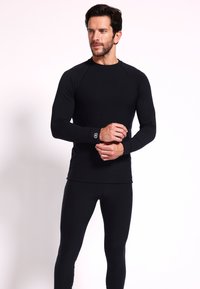 Schwarzes Langarmshirt mit einem figurbetonten Design und gerippter Textur, kombiniert mit schwarzen, figurbetonten Leggings. Verfügt über ein Logo am Handgelenk.