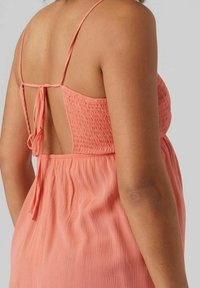 Abito corallo rosa con sottili spalline, dettagli arricciati sulla schiena e scollo basso. Tessuto leggero e testurizzato che fluisce delicatamente.