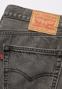 Grå denimjeans med en läderlogga, kontrasterande sömmar och en liten röd etikett på bakfickan. Lös passform, rak stil.