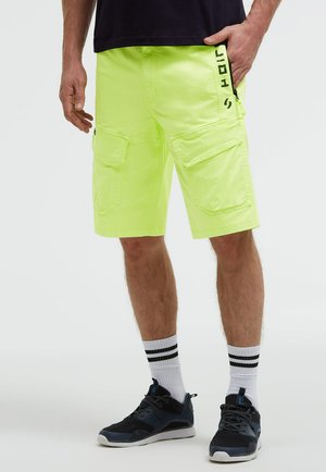 Mann trägt hellgelbe Cargoshorts, schwarze Sneakers, weiße Socken mit schwarzen Streifen und ein schwarzes Shirt, steht mit einer Hand in der Tasche.