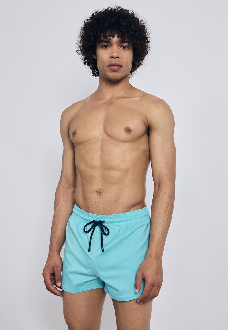 Vīrietis bez krekliem ar cirtainiem matiem, ģērbies gaiši zilos peldkostīmos ar melnu auklu, stāv pret vienkāršu gaiši pelēku fona.