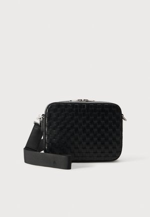 Sac bandoulière rectangulaire noir en matière tissée, avec une large sangle réglable, des accessoires métalliques argentés et une fermeture éclair sur le dessus.