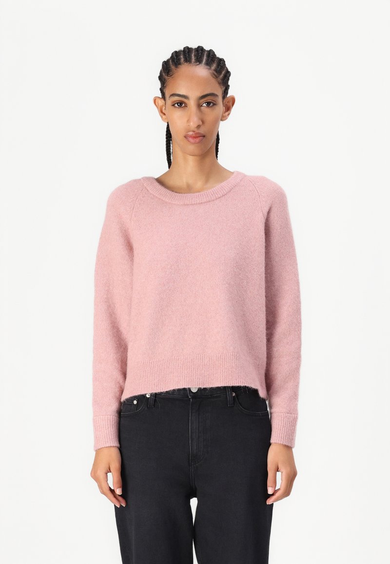 Lys rosa, løs passende strikkegenser med rund hals og ribbestrikkede mansjetter, med en kort lengde og myk tekstur. Bært med svarte jeans.