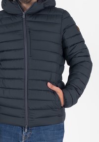 Uomo che indossa un piumino scuro con cappuccio, con una mano nella tasca laterale, abbinato a jeans blu, su uno sfondo neutro.