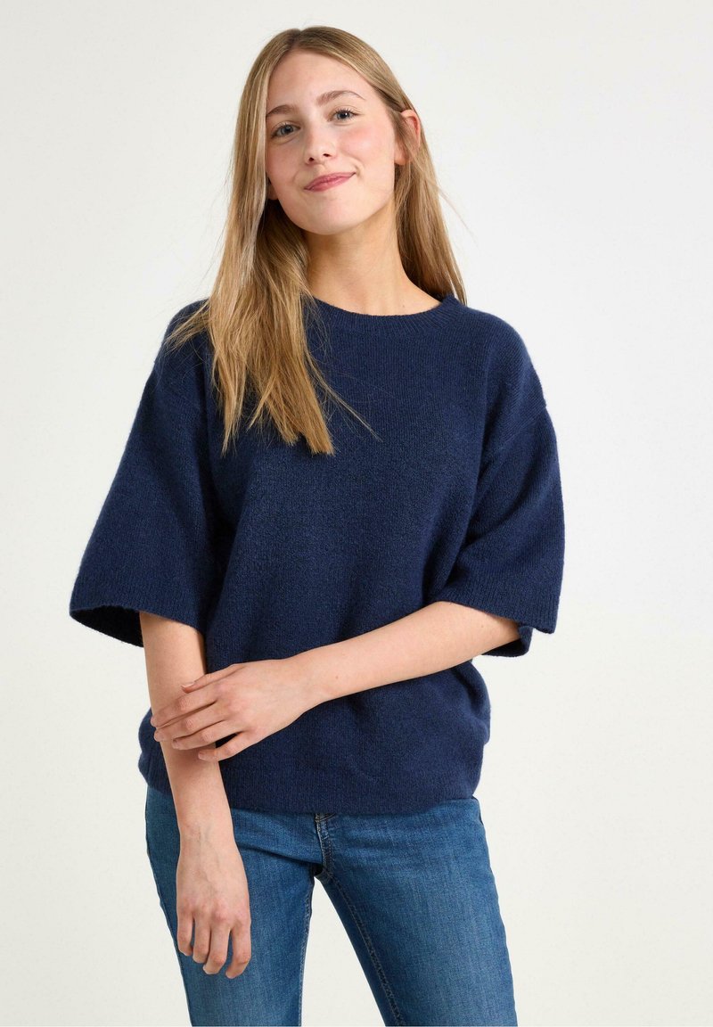 Granatowy sweter dzianinowy z szerokimi krótkimi rękawami, okrągłym dekoltem i miękką fakturą, zestawiony z niebieskimi dżinsami.