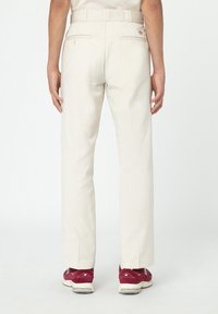 Mann in beigen, geraden Hosen und roten Sneakern, der mit entspannten Händen an den Seiten steht, von hinten fotografiert.