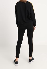 Sweat-shirt noir avec des bandes latérales jaunes, associé à des leggings noirs. Les baskets ont une semelle blanche et un design à lacets noirs.