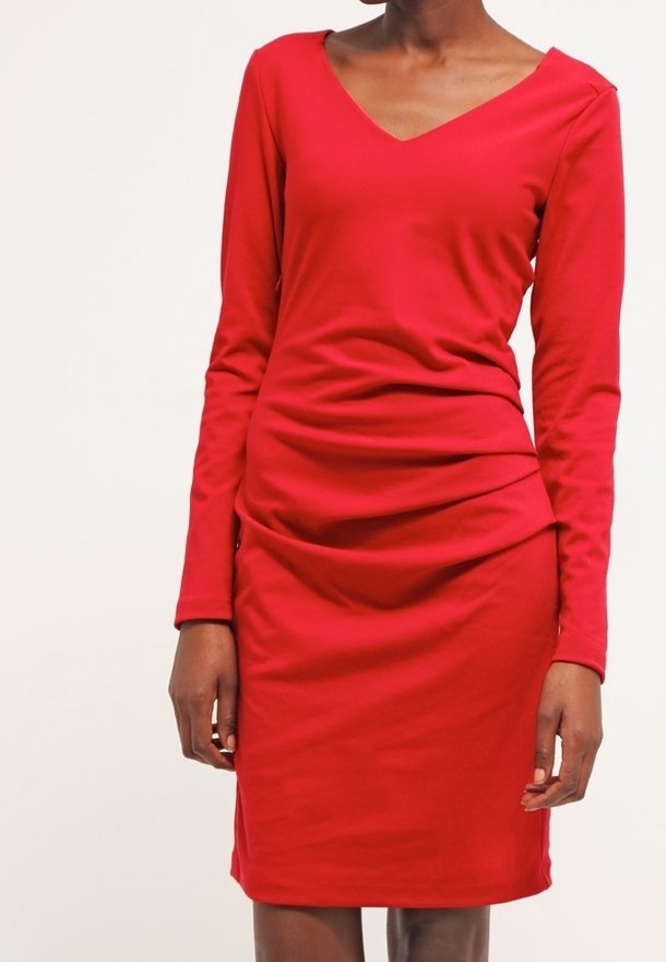 Femme portant une robe rouge ajustée à manches longues, avec un col en V et un léger froncé à l'avant sur un fond clair.