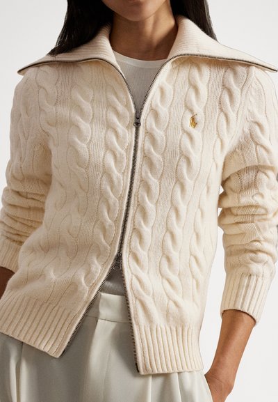 Kardigan z vzorcem pletenja kabel, v krem barvi, z visokim ovratnikom in zadrgo. Rebrasti rokavi in spodnji rob dodajajo strukturo. Zlata podrobnost z logotipom na prsih.