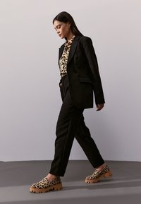 Schwarzer Blazer über einer Bluse mit Leopardenmuster, kombiniert mit schwarzen Hosen und Leopardenmuster-Slippern mit dicken, braunen Sohlen.