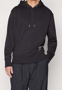 Osoba oblečená v černé mikině s kapucí a šňůrkami a černých kalhotách, jednou rukou v kapse, stojící před světlým pozadím.