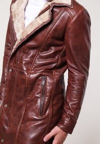 Giubbotto in pelle marrone con colletto in pelliccia, tasca laterale con zip e bottoni a pressione, indossato sopra una camicia bianca.
