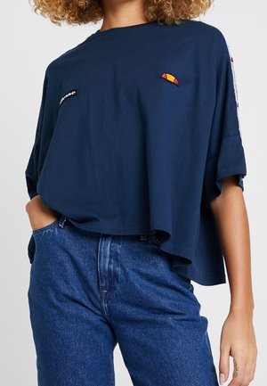 Mujer con camiseta corta azul marino suelta con pequeños logos coloridos, combinada con vaqueros altos azules, mano en el bolsillo.