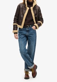 Personne portant une veste en peau lainée marron foncé, un pull en tricot marron foncé, un jean bleu retroussé et des bottes  à lacets marron, debout avec une main dans la poche.