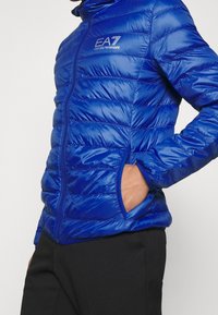 Blauer Puffer-Jacke mit Reißverschluss, horizontalem Steppmuster, Stehkragen und seitlichen Taschen. Mit dem EA7-Logo auf der Brust.