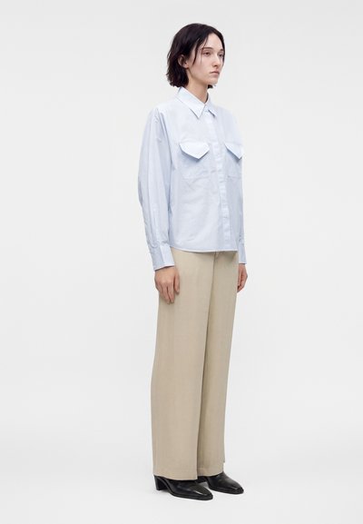 Jeune personne debout portant une chemise bleu pâle à boutons avec des poches poitrine, un pantalon beige à jambes larges, et des chaussures noires à talons, tourné vers la droite.