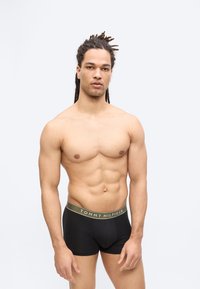 Άνδρας χωρίς πουκάμισο με κοτσίδες, φορώντας μαύρα εσώρουχα Tommy Hilfiger, στέκεται μπροστά σε απλό ανοικτό φόντο.