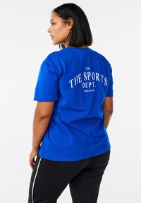 Modrá bavlněná tričko s bílým textem "THE SPORTS DEPT." na zádech, krátké rukávy a volný střih, kombinované s černými kalhotami.