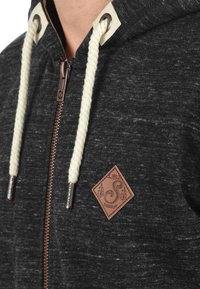 Dark gray hoodie featuring a zip closure, textured fabric, cream drawstrings, and a brown leather logo patch. Copper zipper accents add detail.