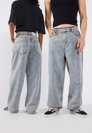 Jeans de mezclilla azul claro con un corte holgado, que cuentan con una cintura alta, cinco bolsillos y dobladillos deshilachados. Combinados con zapatillas negras.