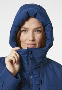 Chaqueta aislante azul con un alto cuello y capucha ajustable. Presenta un cierre frontal con cremallera, cordones en la capucha y un acabado liso y mate.