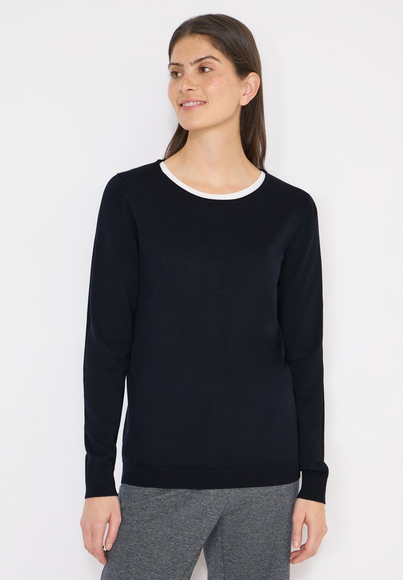 Femme aux longs cheveux foncés portant un pull noir avec un col blanc et un pantalon gris, se tenant devant un fond blanc.