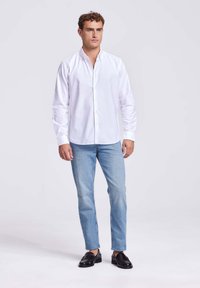 Man staat in een witte blouse met lange mouwen, lichte spijkerbroek en zwarte instappers voor een effen achtergrond.