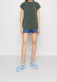 Top verde a maniche corte con scollo arricciato, abbinato a shorts in denim. Sandali leggeri blu chiaro con tacco largo e design traforato.