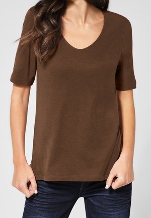 Kvinde iført en brun V-udskærings kortærmet top og mørkeblå jeans, stående med hænderne nær lommerne mod en ensfarvet baggrund.