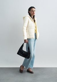 Giacca puffer trapuntata color crema con cappuccio, maglietta gialla, jeans azzurri chiari, borsa nera e scarpe marroni con punta.